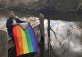 Person holding the Progress Pride Flag outdoors by a river, demonstrating the 3 ft x 5 ft size