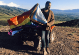 Person displaying the Progress Pride Flag on top of an ATV in a mountain landscape